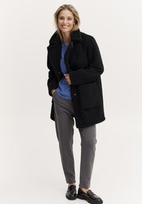 Palton negru din fleece cu guler și buzunare mari, purtat cu o cămașă albastră, pantaloni gri și pantofi negri. Design casual, texturat.
