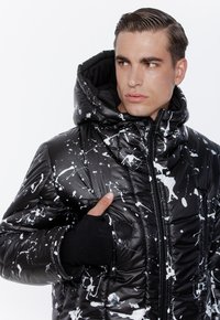 Młody mężczyzna w czarnej kurtce puchowej w wzór w białe plamy farby, z ręką w kieszeni, patrzy w prawo na jednolitym tle.