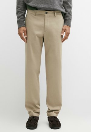 Uomo che indossa pantaloni beige a gamba dritta, maglione grigio e scarpe stringate marrone scuro, in piedi davanti a uno sfondo semplice.