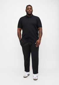 Sort polo-shirt med korte ærmer og en tekstureret finish, parret med løse sorte bukser og hvide sneakers med grå detaljer.
