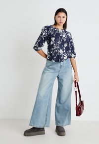 Blouse florale navy à manches bouffantes, associée à un jean large bleu clair et des bottes à plateforme noires. Porte un petit sac bordeaux.