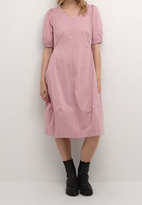 Kvinna klädd i en knälång rosa klänning med puffärmar och V-ringning, i kombination med svarta ankelskor, som står framför en enfärgad vit bakgrund.