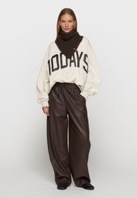 Beige Oversize-Sweatshirt mit schwarzem "100DAYS"-Druck, kombiniert mit braunen, weiten Hosen aus Kunstleder und einem braunen Strick-Schal.