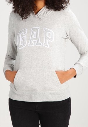 Personne portant un sweat à capuche gris clair avec l'inscription "GAP", mains dans la poche avant, associée à un pantalon noir sur fond blanc.