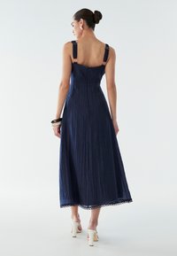 Vestido midi azul marino con tela texturizada, tirantes anchos con detalles decorativos y ribete de encaje en el dobladillo. Combinado con zapatos de tacón blancos.