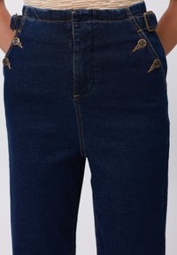 Donkerblauwe denimbroek met een hoge taille, twee decoratieve knopen aan elke kant, contrasterende oranje stiksels en een riemlus.