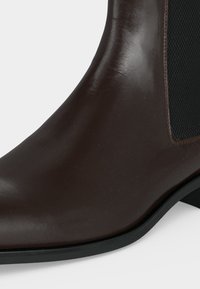Gros plan d'une bottine en cuir marron foncé avec un panneau latéral élastique noir et un petit talon sur fond blanc.