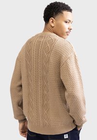 Beige strikket sweater med teksturerede mønstre, et kabelstrikdesign på ryggen og ribbede ærmer og kant.