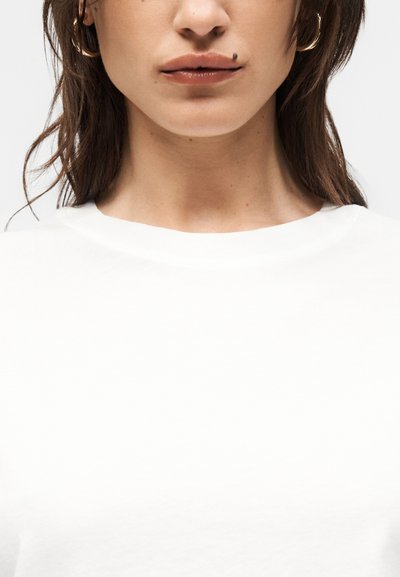 Γυναίκα με καστανά μαλλιά μέχρι τους ώμους που φοράει λευκό πουκάμισο, δείχνοντας τα χείλη της και ένα εμφανές ελιά πάνω από το άνω χείλος στη δεξιά πλευρά.