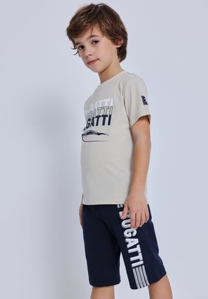 Ragazzo giovane in piedi di lato che indossa una maglietta beige e pantaloncini blu navy, entrambi con la scritta "GATTI" e una grafica di auto sulla maglietta.