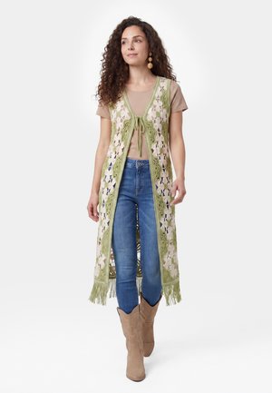 Kvinde med krøllet hår har en beige kortærmet top, lang grøn og cremefarvet hæklevest, blå jeans, brune cowboystøvler og store runde øreringe på.