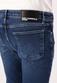 Tamno plave traperice s izblijeđenom teksturom, dizajnom s pet džepova i oznakom na crnom kožnom remenu s logotipom "KARL LAGERFELD JEANS".