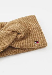 Knit headband in light brown with a twisted design and ribbed texture. Features a small logo patch in red, white, and blue.