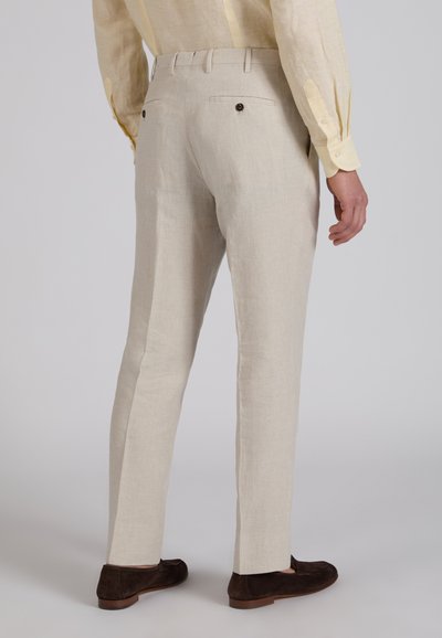 Pantalon beige sur mesure avec poches boutonnées à l'arrière porté par une personne en chemise jaune clair et mocassins marron foncé.