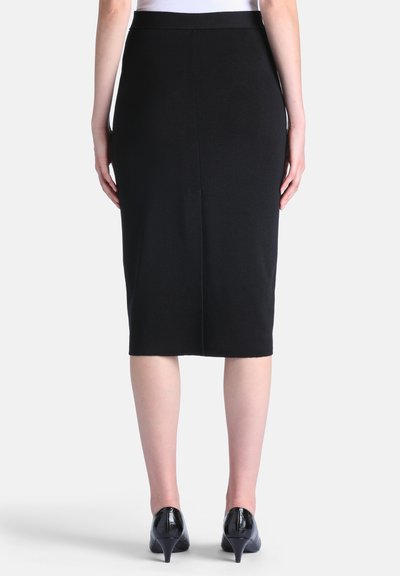 Femme portant une jupe crayon noire ajustée longueur genou et des chaussures noires à talons bas, vue de dos sur un fond uni.