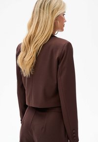 Brązowa krótka marynarka z wyraźnymi ramionami i długimi rękawami. Gładki, dopasowany materiał z detalami na zapięciach rękawów. Prosty, nowoczesny design.