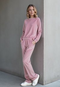 Világos rózsaszín, puha anyagú lounge szett, amely tartalmaz egy kerek nyakú pulóvert és hozzá illő nadrágot zsebekkel; fehér tornacipővel párosítva.