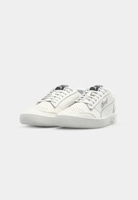 Hvite lær sneakers med perforerte detaljer, rund tå, flat gummisåle, og svarte logo aksenter på tungen og siden. Snøring i matchende farge.