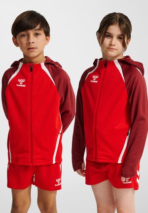 Røde atletiske jakker med hætte og hvide detaljer, parret med matchende røde shorts. Begge beklædningsgenstande har Hummel-logoer og et strømlinet design.