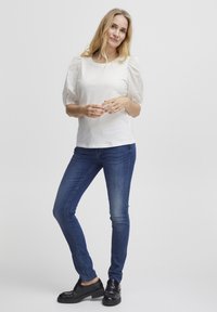 Hvid top med strukturerede puffærmer, parret med blå skinny jeans og sorte sko. Ærmerne har detaljerede øje-mønstre.