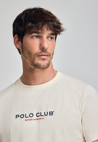 Beżowa koszulka z krótkim rękawem z okrągłym dekoltem, z napisem "POLO CLUB" w kolorze granatowym i "ESTABLISHED 2012" w kolorze czerwonym. Gładka bawełniana struktura.