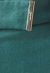 Teal stoff med en myk tekstur, med en gullmetallakzent med gravert logo. Designet inkluderer en brettet lukking.