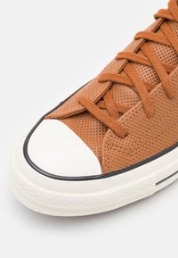 Basket en cuir perforé marron avec capuchon en caoutchouc blanc, contour noir contrastant et lacets marron assortis. Détail supérieur texturé.