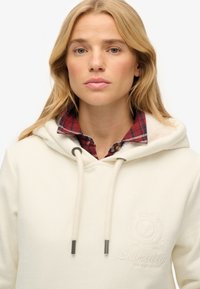 Sudadera blanca con cordón, logo bordado, textura suave, combinada con una camisa de cuadros roja y negra debajo.