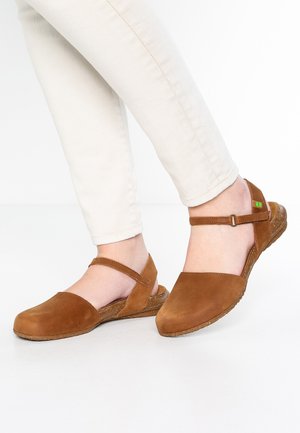 Chaussures en cuir marron à enfiler avec une sangle, surface texturée et semelle en liège, associées à un pantalon slim clair.
