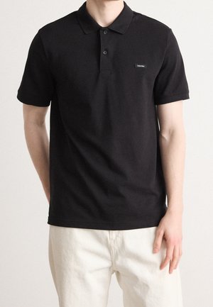 Mężczyzna ubrany w czarną koszulkę polo z krótkim rękawem, z dwoma guzikami i małym logo Calvin Klein, połączoną z białymi spodniami, stoi na jednolitym tle.