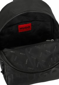 Zaino nero con una finitura liscia e opaca. L'interno presenta un motivo con logo e un'etichetta "HUGO" rossa ben visibile. Chiusura con zip.