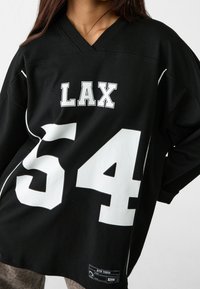 Maillot de sport noir oversize avec accents blancs, arborant le texte "LAX" et le numéro 54 en grand. Fabriqué en tissu doux avec un col en V.