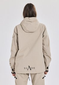 Beige wetterfeste Jacke mit Kapuze, mit einem Logo auf dem Rücken, verstellbaren Bündchen und einer glatten, matten Textur.