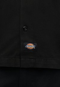 Fekete szövet gombbal és egy téglalap alakú, narancs, piros és fehér színekben a Dickies logóját viselő felvarróval.