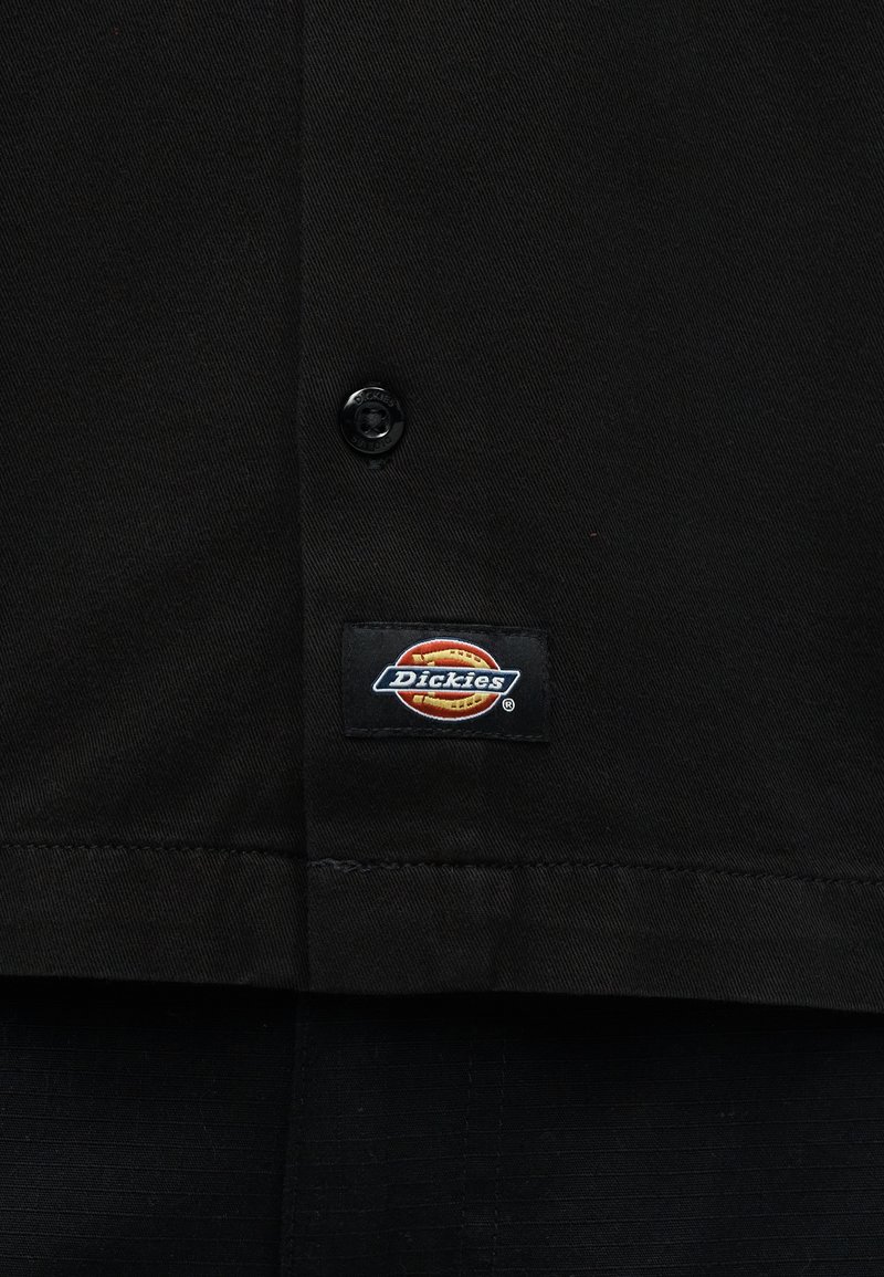 Tessuto nero con un bottone e un'etichetta rettangolare cucita con il logo Dickies nei colori arancione, rosso e bianco.