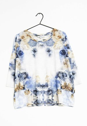 Blusa suelta de manga larga con un estampado floral en azul, beige y blanco, colgada en una percha negra contra una pared blanca.