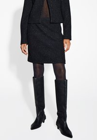 Schwarze strukturierte Anzugjacke und Rock mit metallischen Einsprengseln, kombiniert mit knielangen schwarzen Lederstiefeln, die ein figurbetontes Design zeigen.