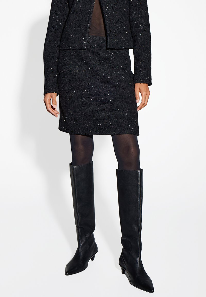 Schwarze strukturierte Anzugjacke und Rock mit metallischen Einsprengseln, kombiniert mit knielangen schwarzen Lederstiefeln, die ein figurbetontes Design zeigen.
