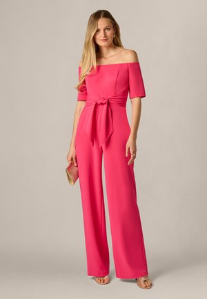 Femme aux longs cheveux blonds portant une combinaison rose vif à épaules dénudées avec une taille nouée, tenant une petite pochette et portant des sandales beiges.