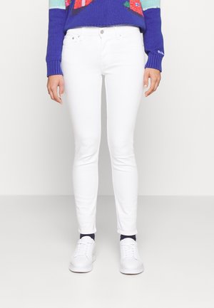 Persoon die nauwsluitende witte broek, witte sneakers en een blauwe trui met kleurrijke patronen draagt, staand tegen een effen achtergrond.