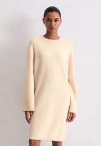 Robe en tricot beige clair, avec un col rond et des manches longues. Texture douce avec une coupe ample, atteignant la mi-cuisse.