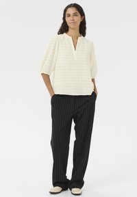 Blusa bianca testurizzata con scollatura a V e maniche a sbuffo. Abbinata a pantaloni neri a righe e scarpe di colore chiaro.