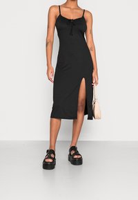 Vestido negro sin mangas con tirantes ajustables, abertura en la parte delantera y detalle de cuerpo fruncido. La modelo lleva sandalias de plataforma y sostiene un bolso pequeño y ligero.
