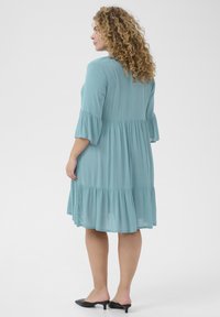 A light teal, knee-length dress in a flowy fabric with a tiered ruffle hem and three-quarter bell sleeves. It features a relaxed fit.