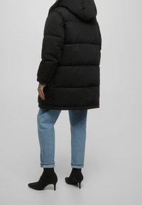 Manteau matelassé noir avec capuche, présentant un design matelassé et des poignets élastiques, associé à un jean bleu clair et des bottines noires à bout pointu.
