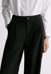 Pantaloni neri su misura realizzati in tessuto testurizzato, con un motivo maculato, chiusura con bottone e tasche laterali. Abbinati a una camicia bianca.