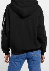 Sudadera negra de gran tamaño con una capucha grande, puños acanalados y un cordón, con una cinta de logo blanca a lo largo de las mangas.