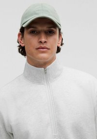 Grauer Reißverschluss-Sweatshirt mit hohem Kragen, das eine glatte Textur aufweist. Hellgrüne Kappe mit gesticktem Branding. Einfaches, minimalistisches Design.