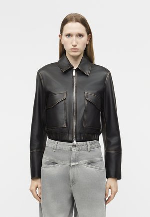 Γυναίκα που φοράει μαύρο cropped δερμάτινο μπουφάν με φερμουάρ μπροστά και δύο μεγάλες μπροστινές τσέπες, συνδυασμένο με γκρι τζιν ψηλόμεσα.