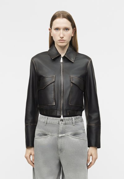 Femme portant une veste en cuir noire courte avec fermeture éclair à l’avant et deux grandes poches à l’avant, associée à un jean gris taille haute.
